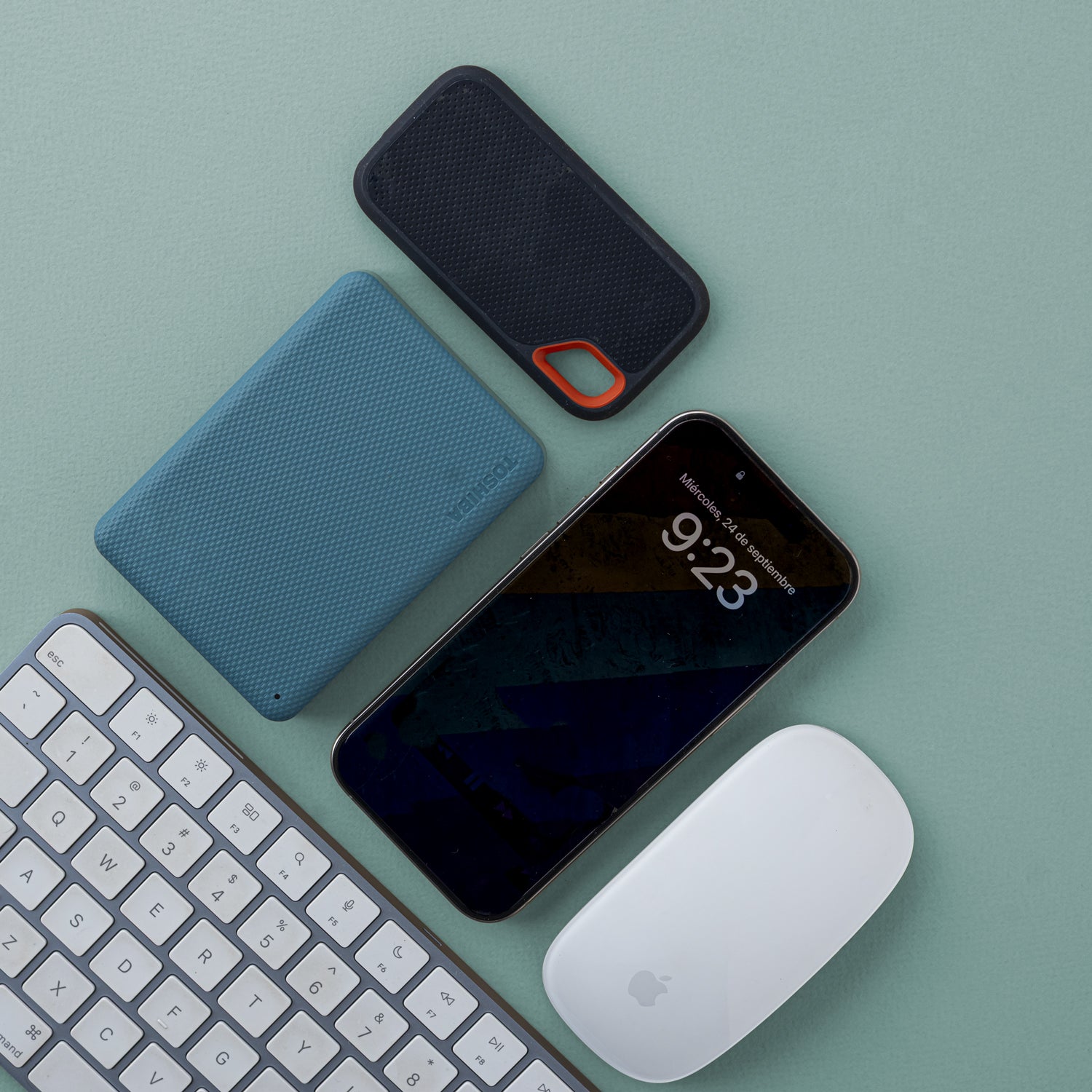 Un celular, un disco duro externo, un mouse inalámbrico y un teclado inalámbrico sobre una mesa.